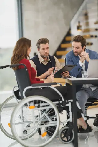 3 people, 2 men and a woman.  The woman is in a wheelchair.  they are at work by a table in conversation